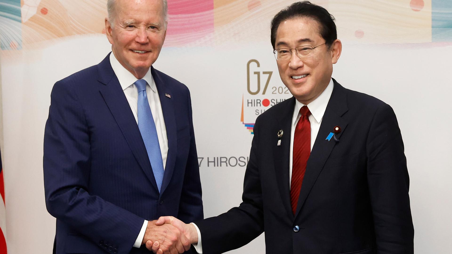 US-Präsident Joe Biden und der japanische Ministerpräsident Fumio Kishida stehen nebeneinander und geben sich die Hand. Sie lächeln in die Kamera. Im Hintergrund ist an einer Wand das Logo des G-7-Treffens zu sehen.