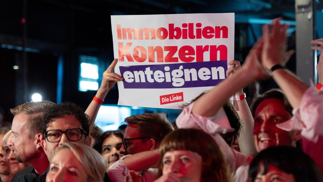 Feiernde Menschen bei der Wahlparty der Partei Die Linke am 20.9.2026. Im Hintergrund hält ein Mitglied ein Plakat mit der Aufschrift "Immobilienkonzern enteignen" hoch.