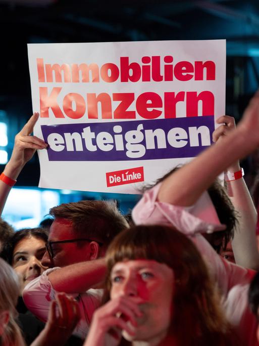 Feiernde Menschen bei der Wahlparty der Partei Die Linke am 20.9.2026. Im Hintergrund hält ein Mitglied ein Plakat mit der Aufschrift "Immobilienkonzern enteignen" hoch.