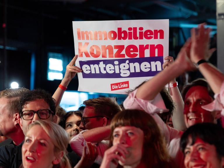 Feiernde Menschen bei der Wahlparty der Partei Die Linke am 20.9.2026. Im Hintergrund hält ein Mitglied ein Plakat mit der Aufschrift "Immobilienkonzern enteignen" hoch.