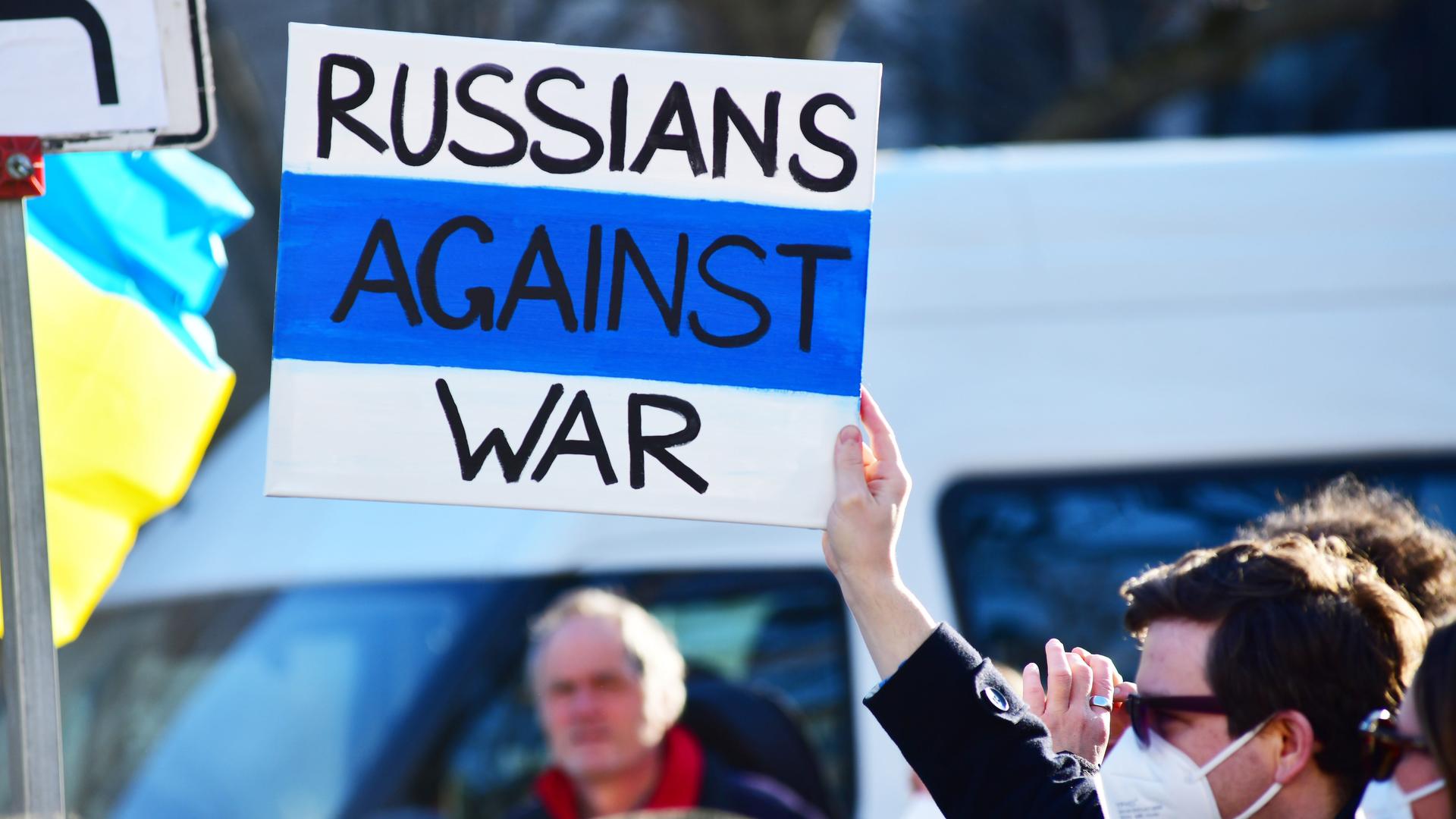 Demonstration in Berlin gegen den russischen Krieg in der Ukraine. Links sieht man eine blau-gelbe Fahne, rechts hält ein Mann ein Plakat mit der Aufschrift "Russians against War" in die Luft.