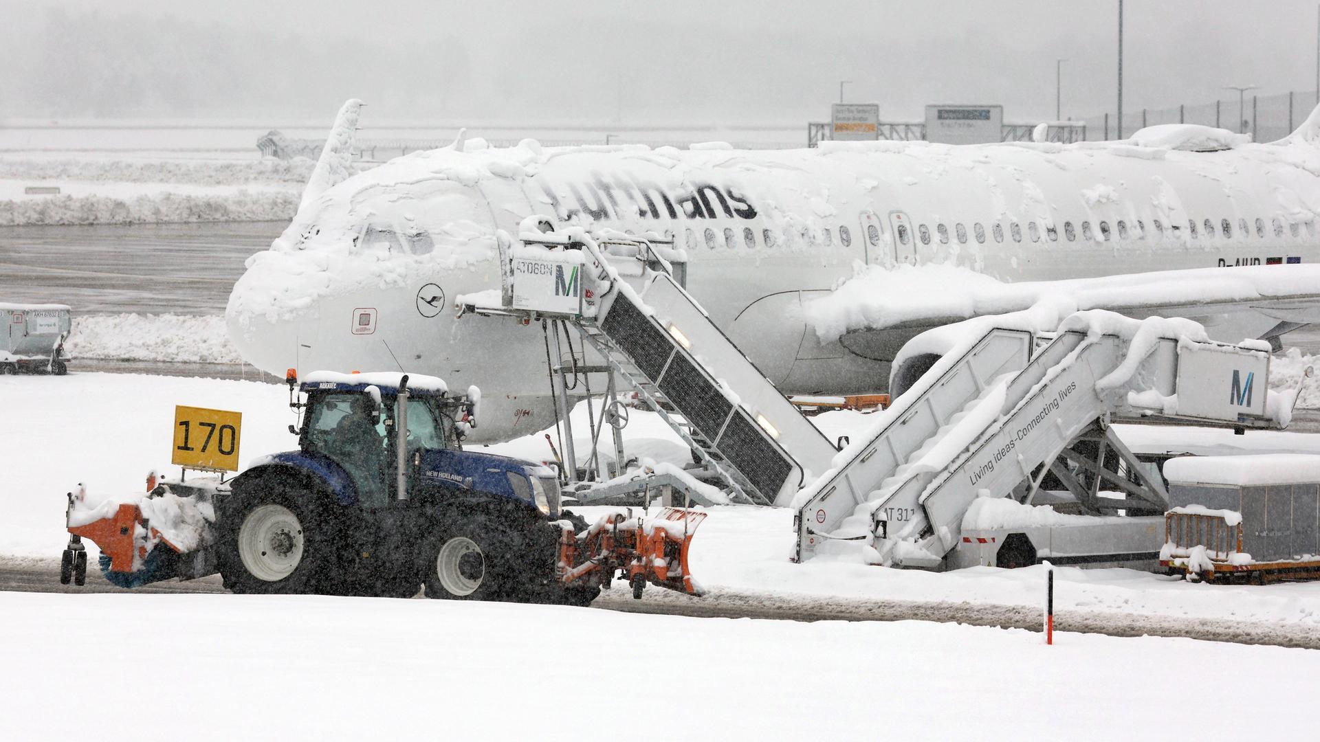 Ein Räum- und Streufahrzeug fährt im Schneetreiben auf dem Flughafen München an einer verschneiten Maschine von Lufthansa vorbei.