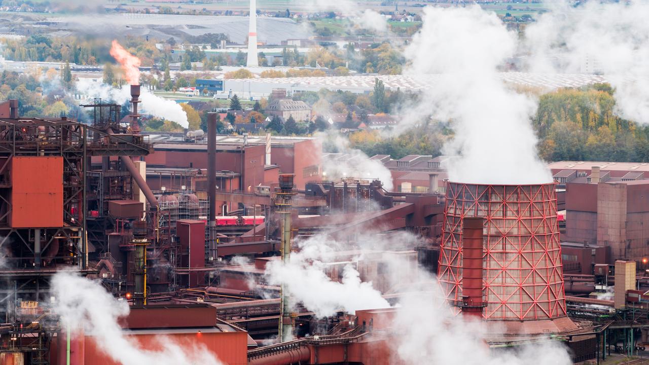 Luftaufnahme eines großen Stahlwerks: rostrot-braune Industrieanlagen, Hochöfen und ein markanter Kühlturm, aus mehreren Schloten steigt dichter weißer Dampf auf. Im Hintergrund liegen Wälder und flaches Land unter bedecktem Himmel.