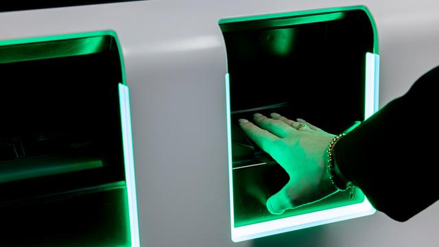 Eine Person legt die Hand in einen Scanner. Der Scanner nimmt die Finger-Abdrücke der Person ab. In dem Scanner leuchtet grünes Licht.