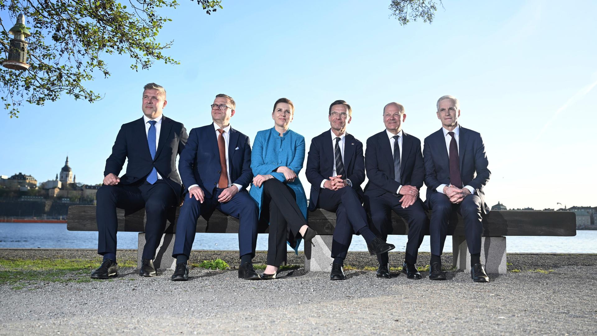 Bundeskanzler Scholz und die Regierungschefs der nordischen Länder sitzen nebeneinander auf einer Bank. Im Hintergrund sind Wasser und blauer Himmel zu sehen. 