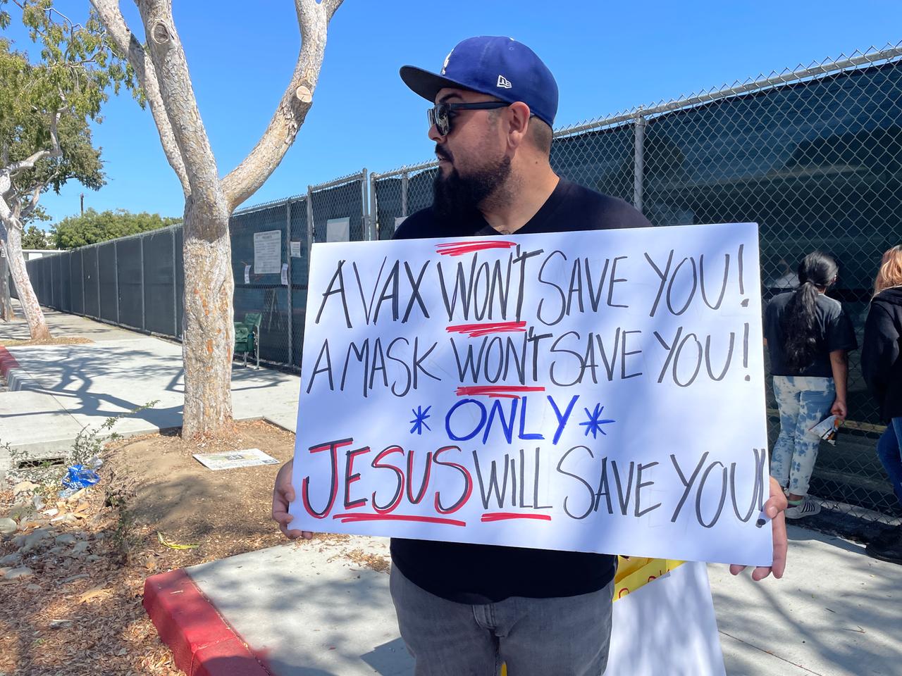 Ein Mann mit Bart und dunkler Sonnenbrille hält auf der Straße ein Plakat hoch, auf dem steht, dass nicht Masken retten, sondern Jesus rettet.