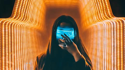 Eine Person im schwarzen Oberteil steht mit einer digitalen Maske in einem orange beleuchteten-Raum.