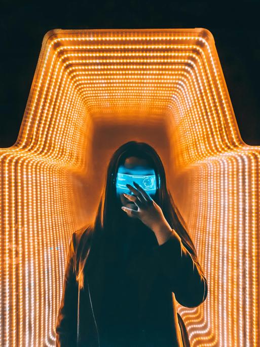 Eine Person im schwarzen Oberteil steht mit einer digitalen Maske in einem orange beleuchteten-Raum.