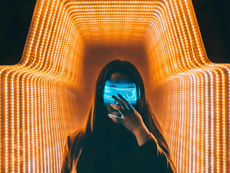 Eine Person im schwarzen Oberteil steht mit einer digitalen Maske in einem orange beleuchteten-Raum.