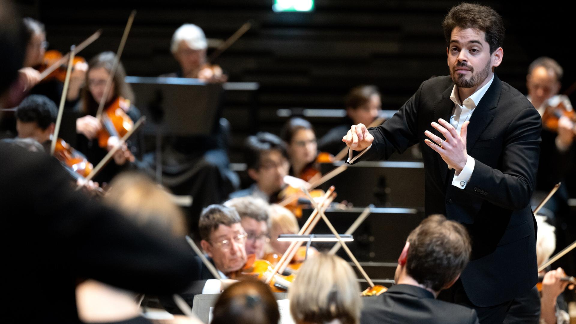 Lahav Shani, zukünftiger Chefdirigent der Münchner Philharmoniker, steht zwischen Musikern auf der Bühne und dirigiert Lahav Shani, zukünftiger Chefdirigent der Münchner Philharmoniker, steht zwischen Musikern auf der Bühne und dirigiert