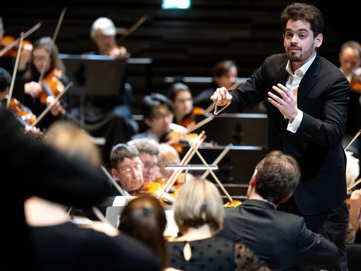 Lahav Shani, zukünftiger Chefdirigent der Münchner Philharmoniker, steht zwischen Musikern auf der Bühne und dirigiert