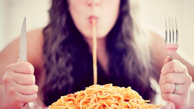 Eine Frau sitzt vor einem Teller Spaghetti und saugt eine Nudel ein, in den Händen hält sie Gabel und Messer