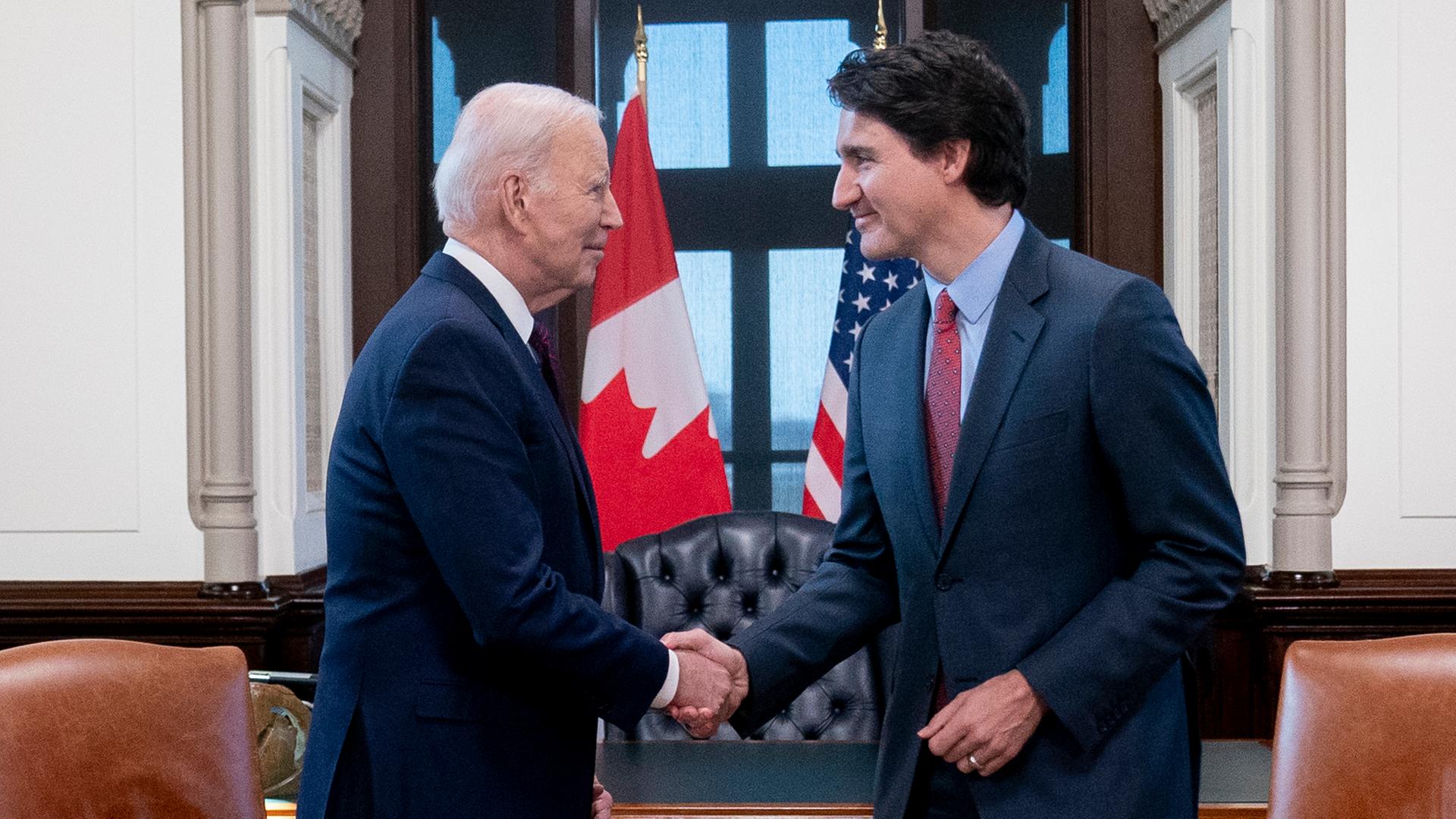 Biden schüttelt dem kanadischen Premierminister Trudeau die Hand. Im Hintergrund sieht man die us-amerikanische und die kanadische Flagge.