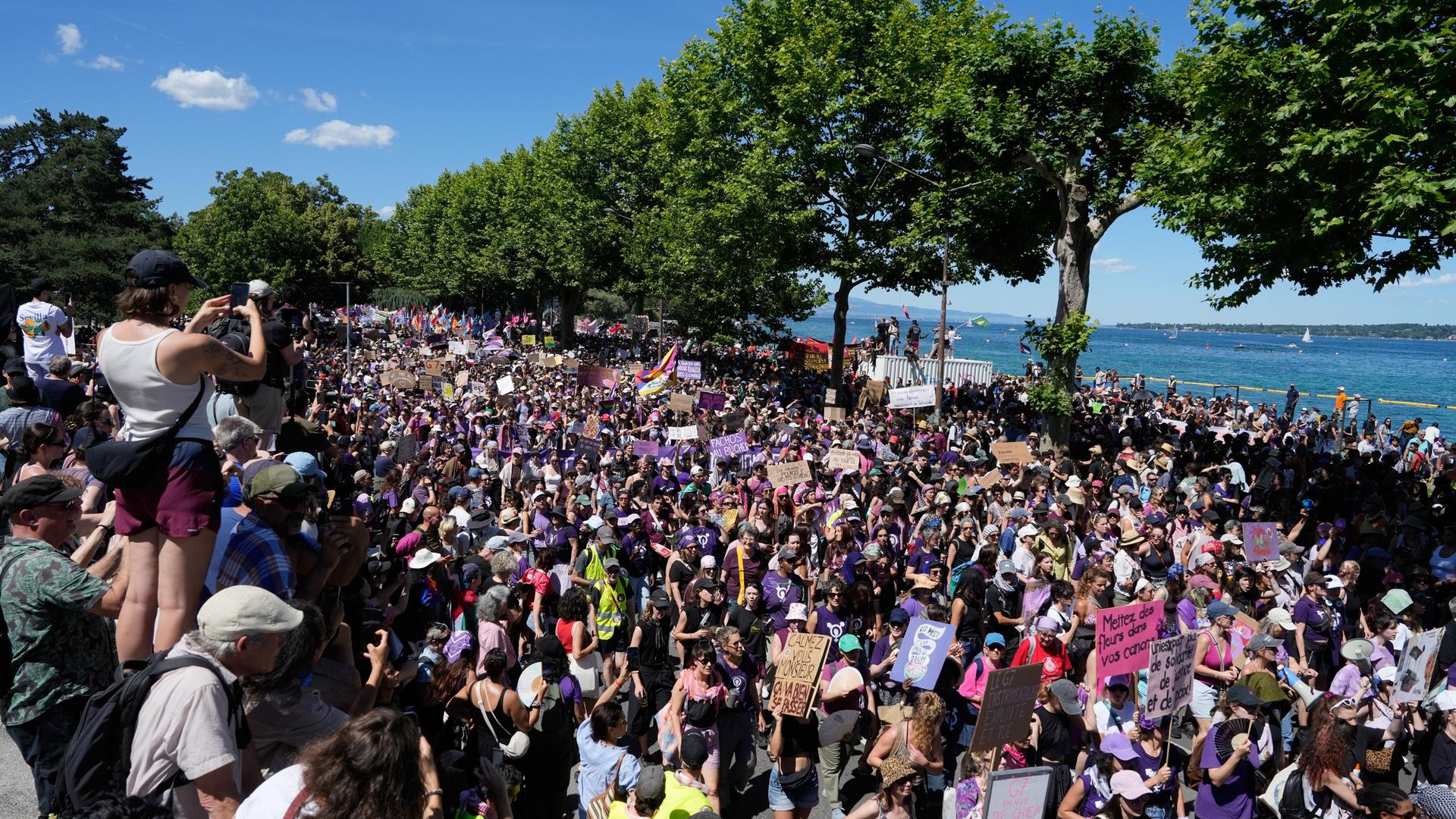 Eine Menschenmenge zieht in Genf am Ufer eines Gewässers entlang. Einige Demonstranten tragen Schilder. Links im Bild beobachten und fotografieren Menschen die Proteste. Eine Menschenmenge zieht in Genf am Ufer eines Gewässers entlang. Einige Demonstranten tragen Schilder. Links im Bild beobachten und fotografieren Menschen die Proteste.