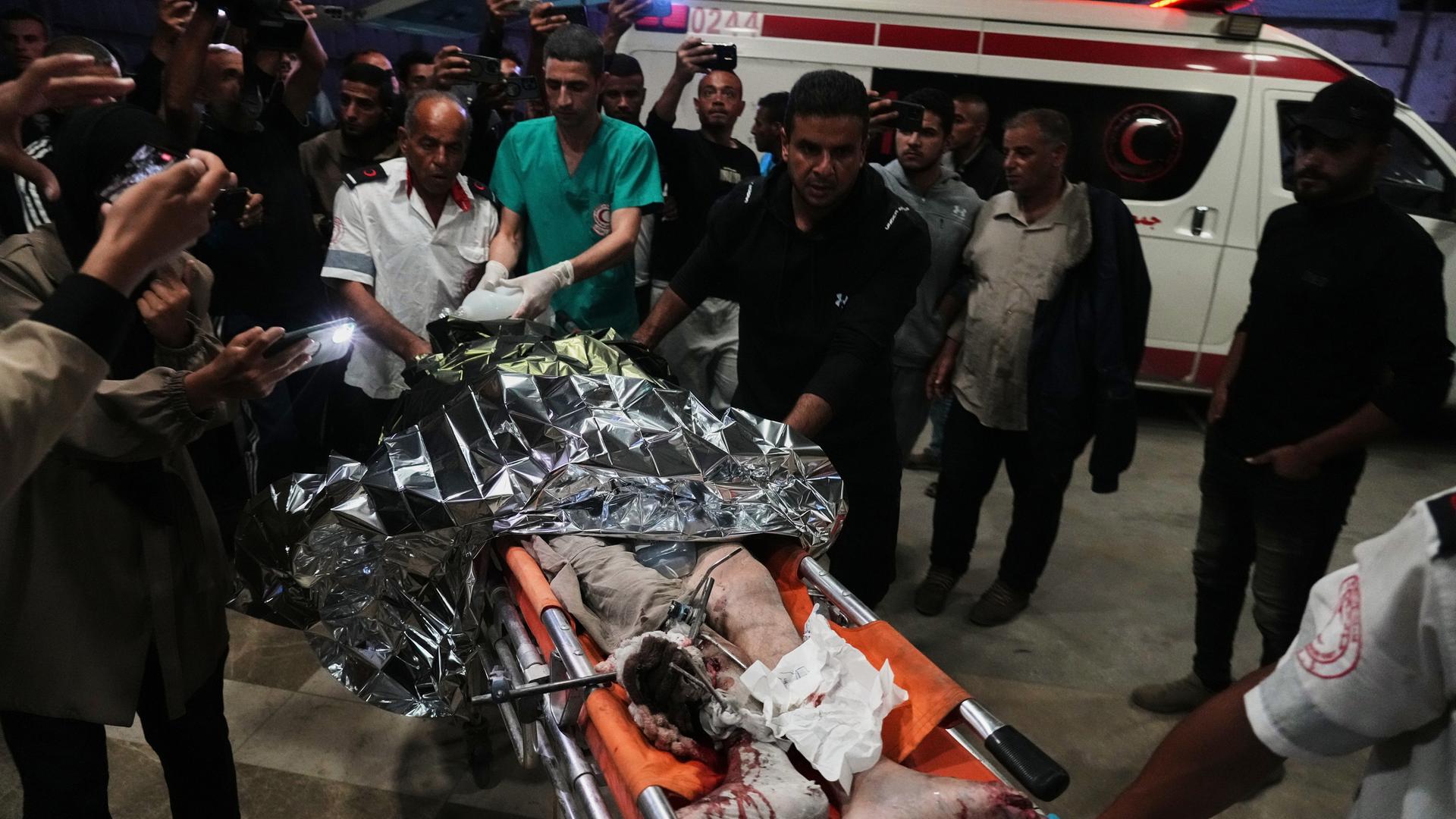 Der Palästinenser liegt auf einer orangefarbenen Bahre, man sieht seine verletzten Beine. Der Oberkörper ist von einer Silberfolie abgedeckt. Sanitäter tragen die Bahre, um sie herum weitere Menschen. Im Hintergrund ein Rettungswagen. Es ist dunkel.  Der Palästinenser liegt auf einer orangefarbenen Bahre, man sieht seine verletzten Beine. Der Oberkörper ist von einer Silberfolie abgedeckt. Sanitäter tragen die Bahre, um sie herum weitere Menschen. Im Hintergrund ein Rettungswagen. Es ist dunkel.