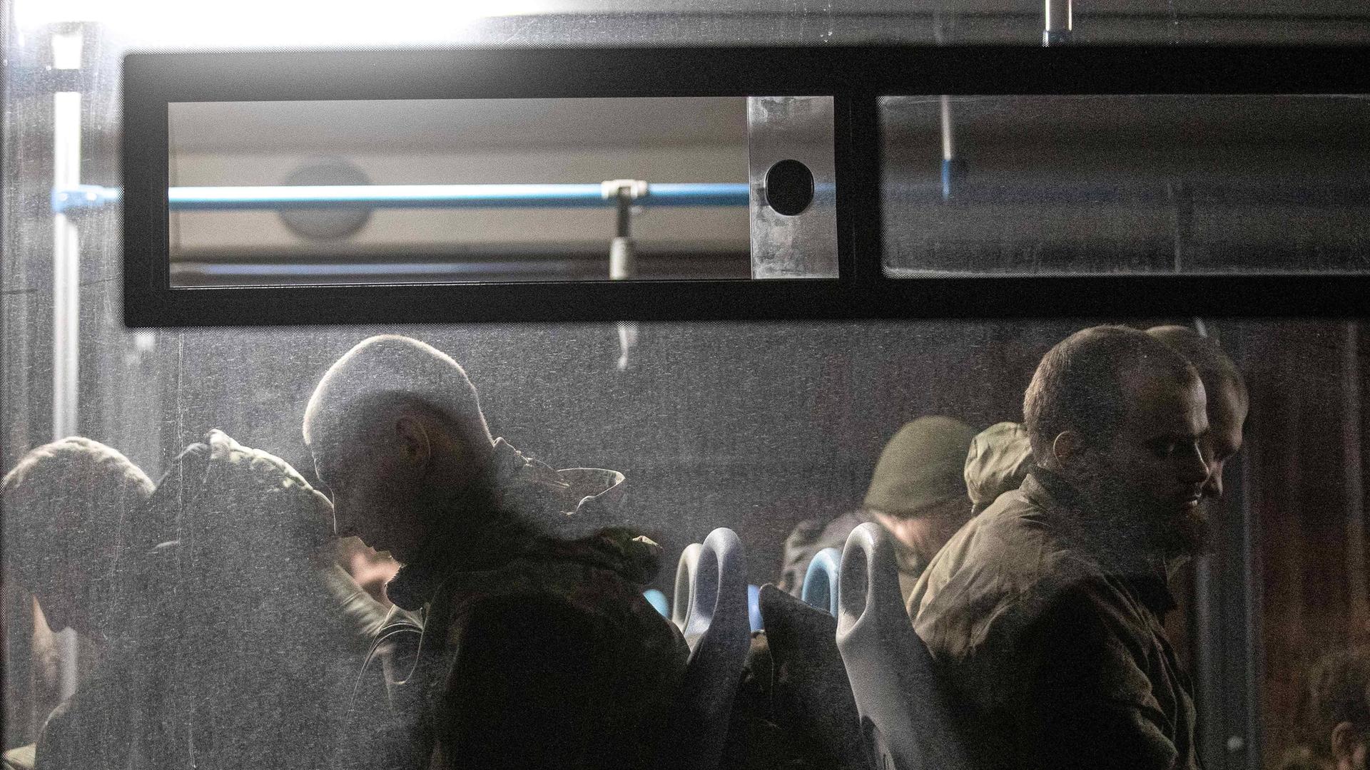 Durch eine Busfensterscheibe sieht man mehrere Soldaten sitzen, die von oben beleuchtet werden. Draußen ist es dunkel.