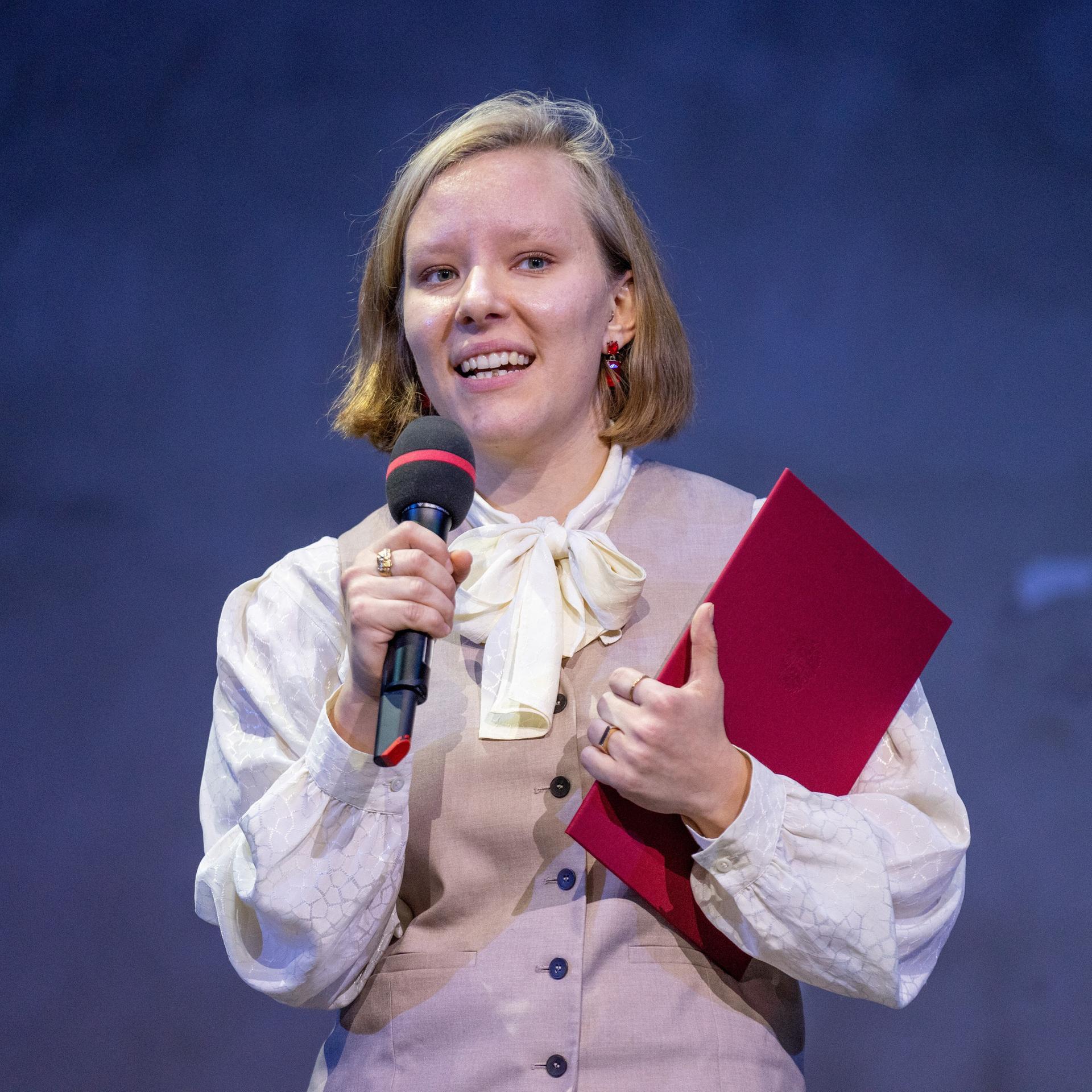 Die Autorin Raphaela Edelbauer hält ein Mikrofon in der einen und ein rotes Buch in der anderen Hand.