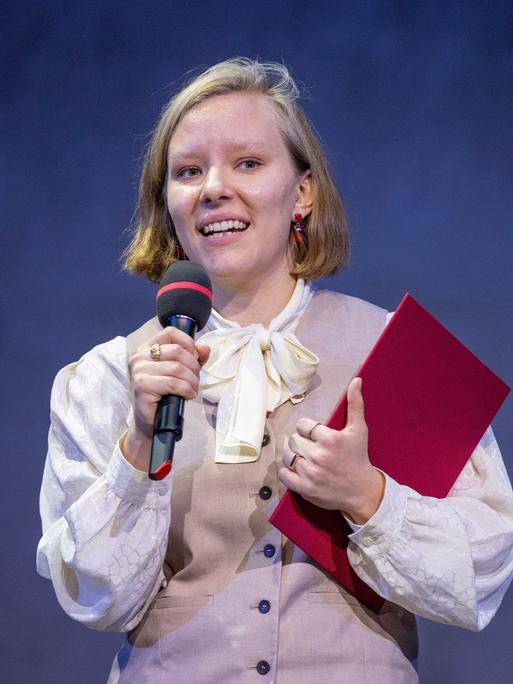 Die Autorin Raphaela Edelbauer hält ein Mikrofon in der einen und ein rotes Buch in der anderen Hand.