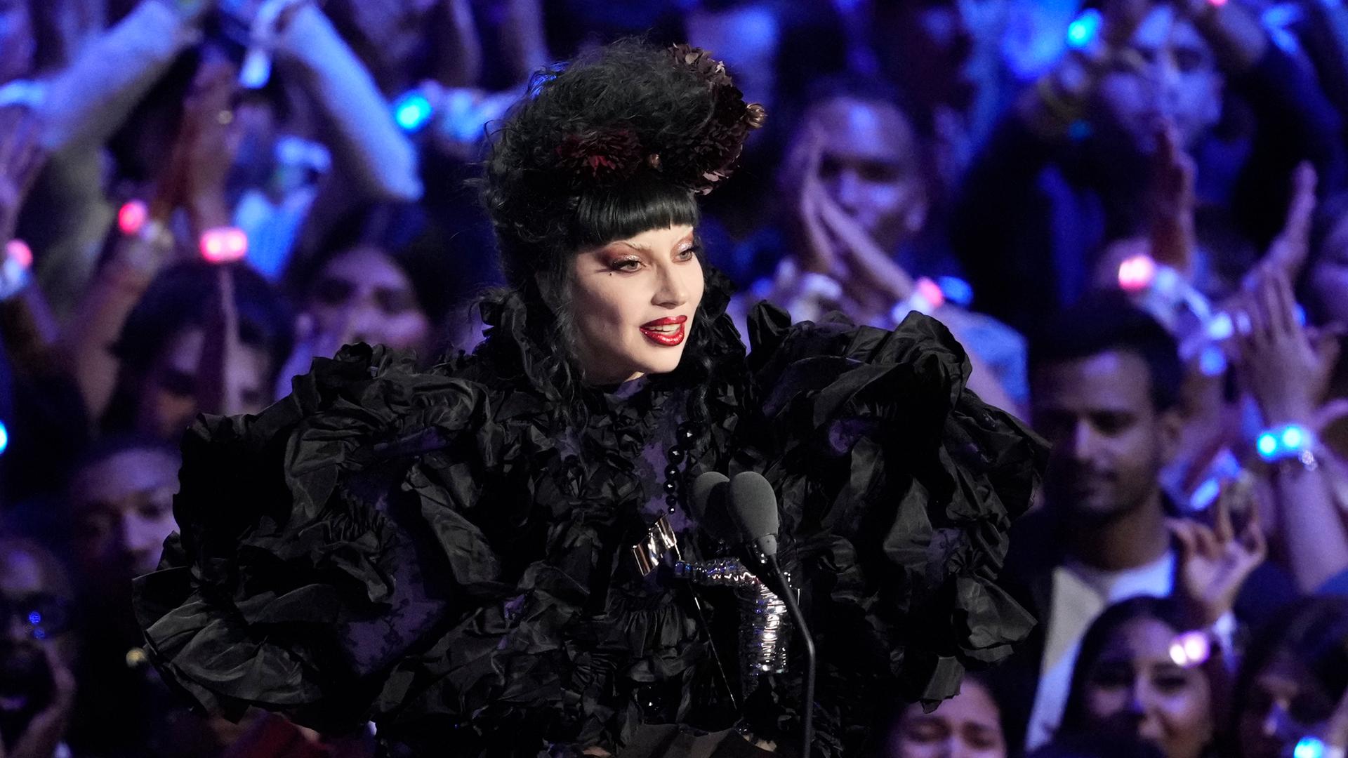 Lady Gaga steht im schwarzen Kostüm vor einem Mikrofon, hinter ihr sind jubelnde Fans zu sehen.