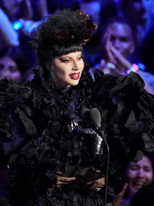 Lady Gaga steht im schwarzen Kostüm vor einem Mikrofon, hinter ihr sind jubelnde Fans zu sehen.