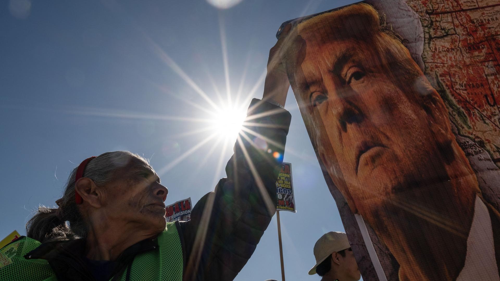 Eine Demonstrantin im Gegenlicht der Sonne hält ein Banner mit dem Gesicht von Donald Trump in die Höhe.