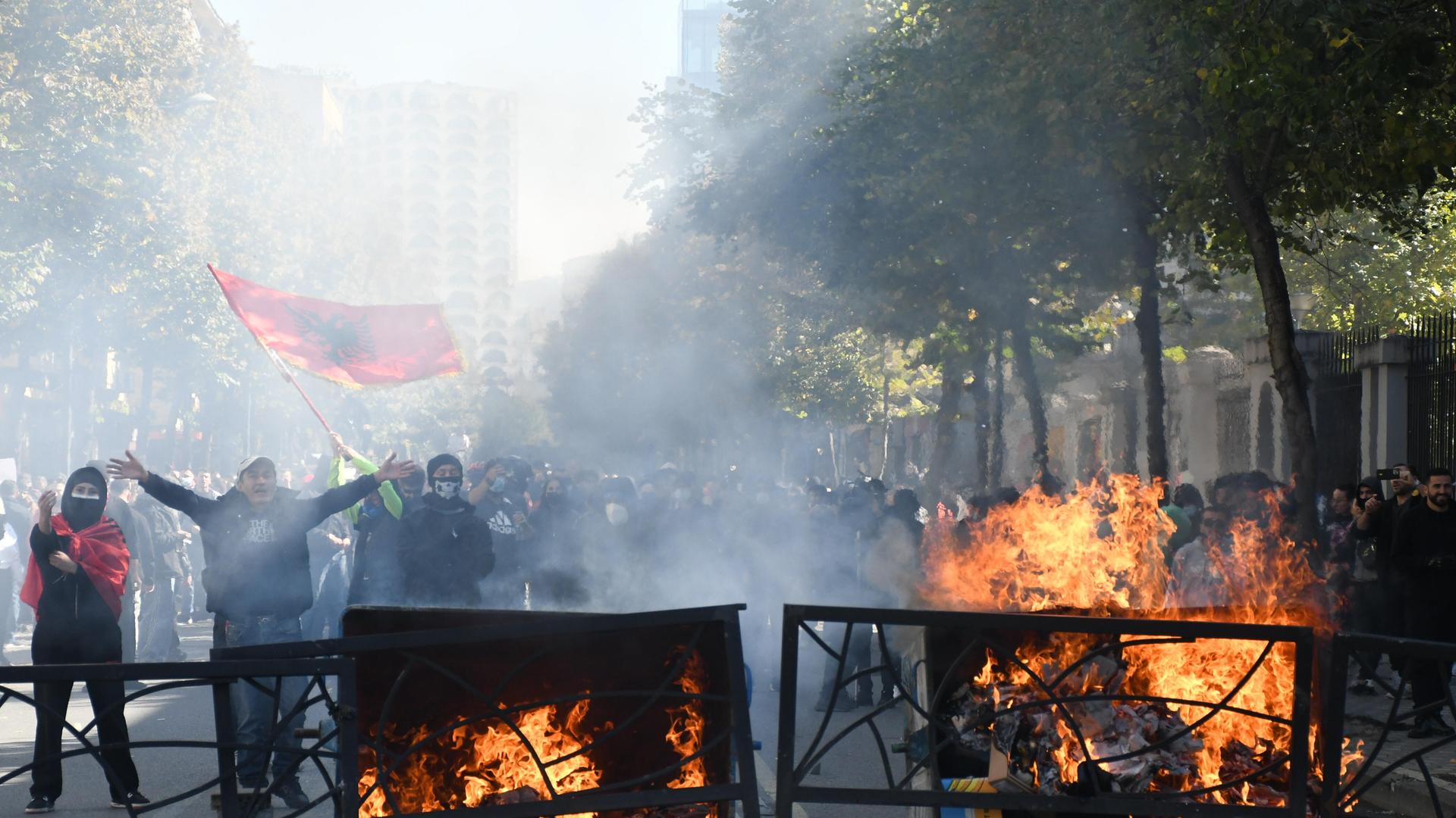 Das Bild zeigt brennende Barrikaden auf einer Straße. Dahinter sind im Rauch vermummte Protestierende zu erkennen. Einer von ihnen schwenkt eine rote Fahne. Das Bild zeigt brennende Barrikaden auf einer Straße. Dahinter sind im Rauch vermummte Protestierende zu erkennen. Einer von ihnen schwenkt eine rote Fahne.