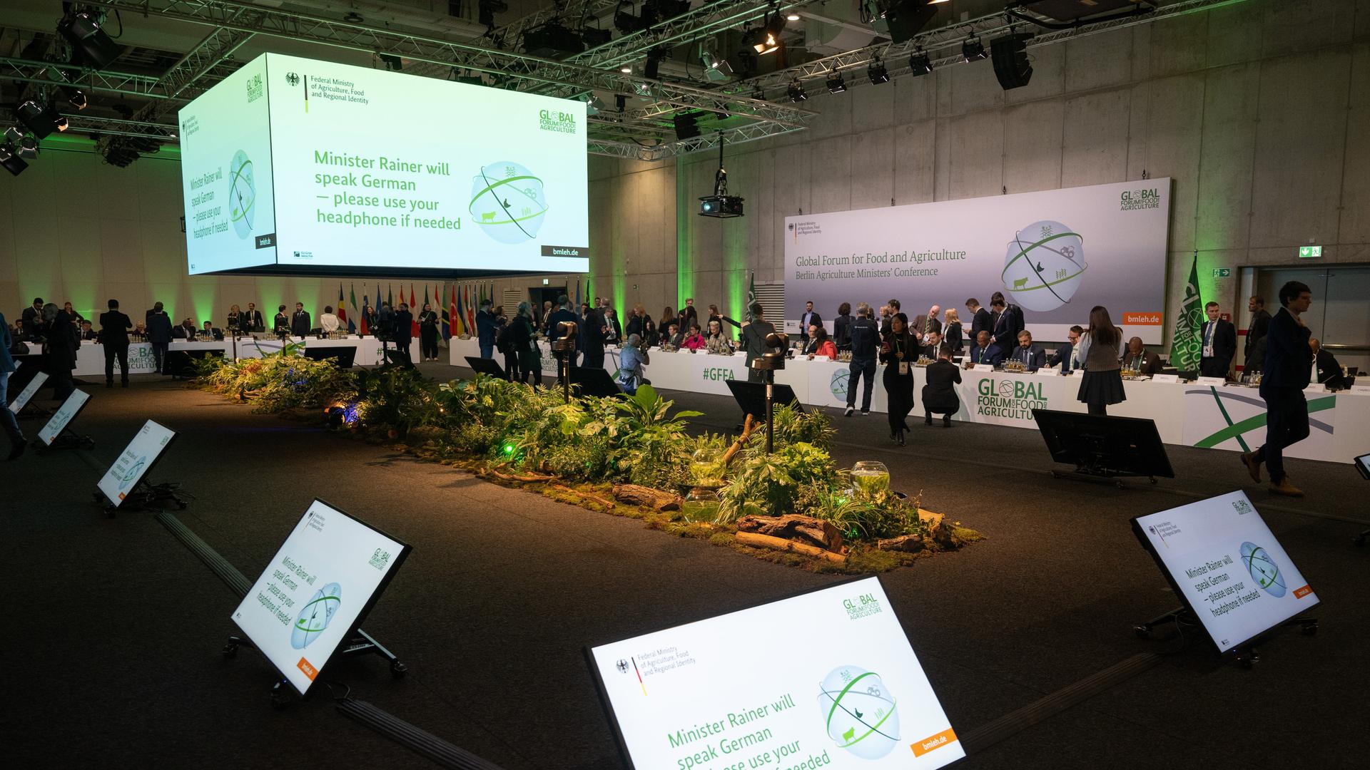 In einem großen Saal stehen mehrere Menschen an langen Tischen, in der Mitte hängt ein großes Plakat, das die Agrarministerkonferenz ankündigt.