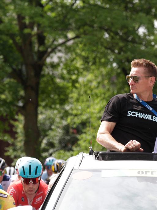 Der Ex-Radsportprofi André Greipel schaut aus dem Dachfenster eines fahrenden Autos auf Radprofis um ihn herum