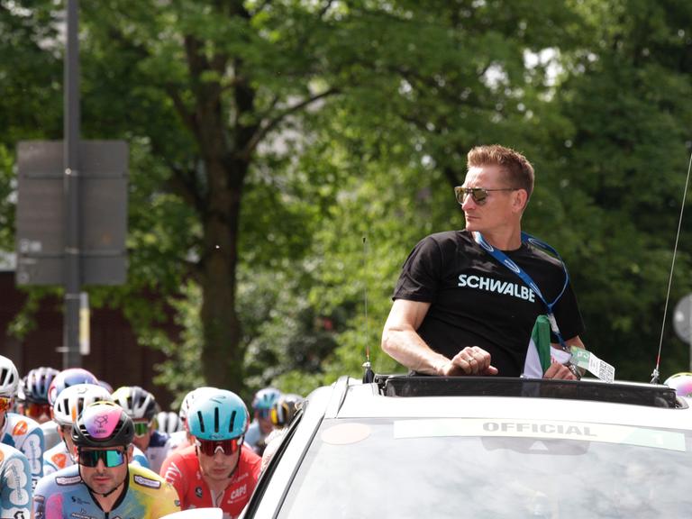 Der Ex-Radsportprofi André Greipel schaut aus dem Dachfenster eines fahrenden Autos auf Radprofis um ihn herum