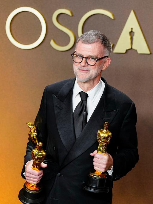 Paul Thomas Anderson, Regisseur des Politthrillers "One Battle After Another" schaut in die Kamera. Er hält zwei der insgesamt sechs Oscars, die der FIlm gewonnen hat, in den Händen.