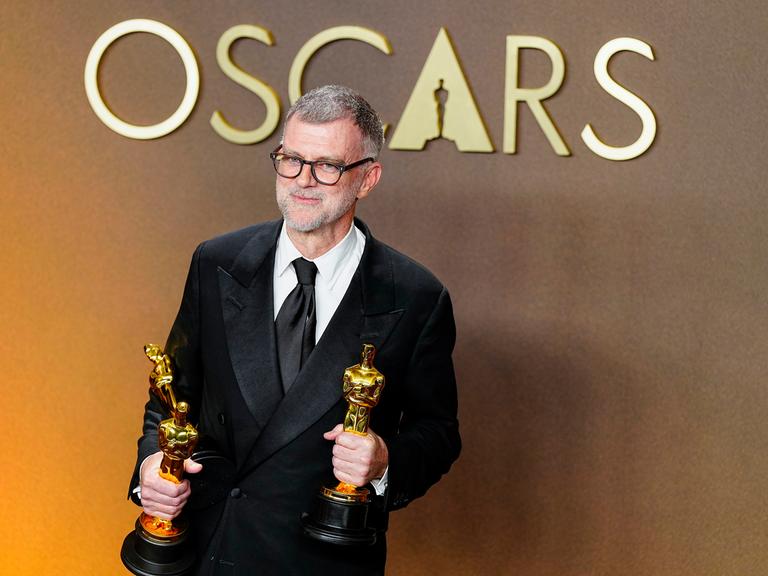 Paul Thomas Anderson, Regisseur des Politthrillers "One Battle After Another" schaut in die Kamera. Er hält zwei der insgesamt sechs Oscars, die der FIlm gewonnen hat, in den Händen.