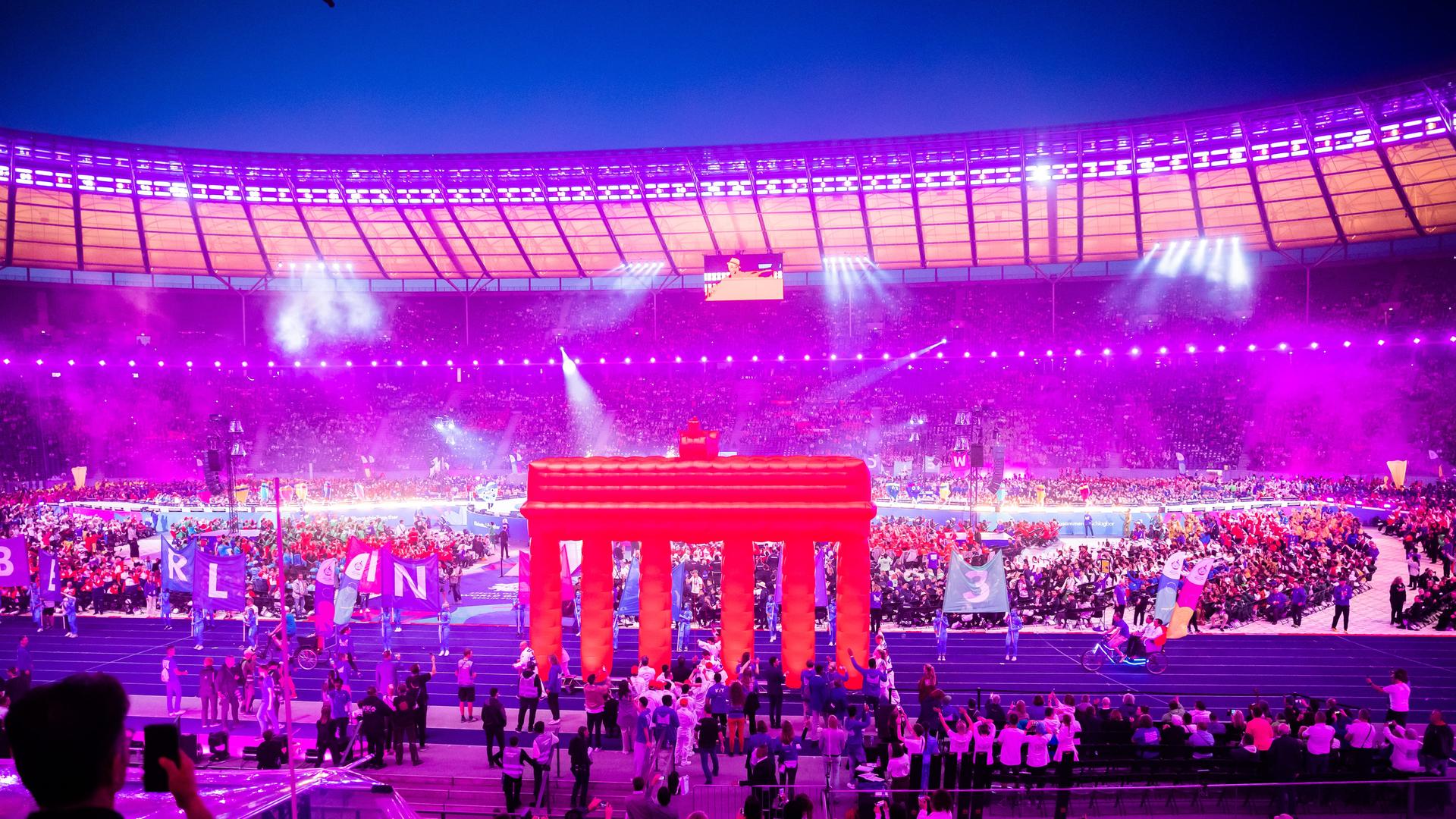 Athleten und Delegationsmitglieder nehmen an der Eröffnungsfeier der Special Olympics World Games Berlin 2023 im Olympiastadion teil. Das Bild ist ein Foto einer Nachtaufnahme des Stadions mit vielen Menschen. Das Brandenburger Tor ist in einer pinkfarbenen Projektion in der Stadionmitte zu sehen.