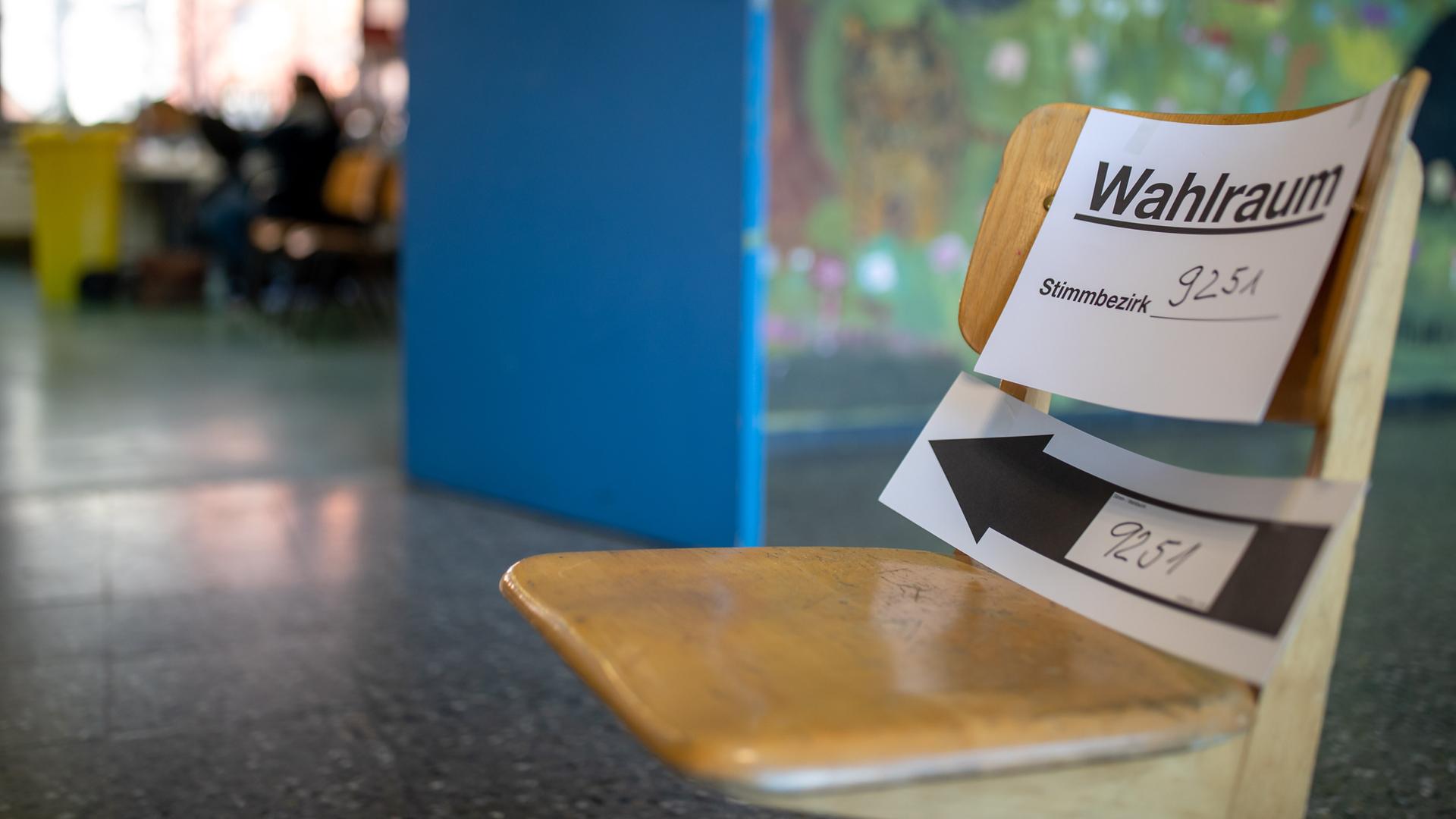 "Wahlraum" steht auf einem Zettel zur Kommunalwahl in Bayern, der in einem Wahllokal an einem Klassenstuhl befestigt ist. Im Hintergrund ist unscharf ein Klassenraum zu erkennen. 