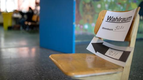 "Wahlraum" steht auf einem Zettel zur Kommunalwahl in Bayern, der in einem Wahllokal an einem Klassenstuhl befestigt ist. Im Hintergrund ist unscharf ein Klassenraum zu erkennen. 