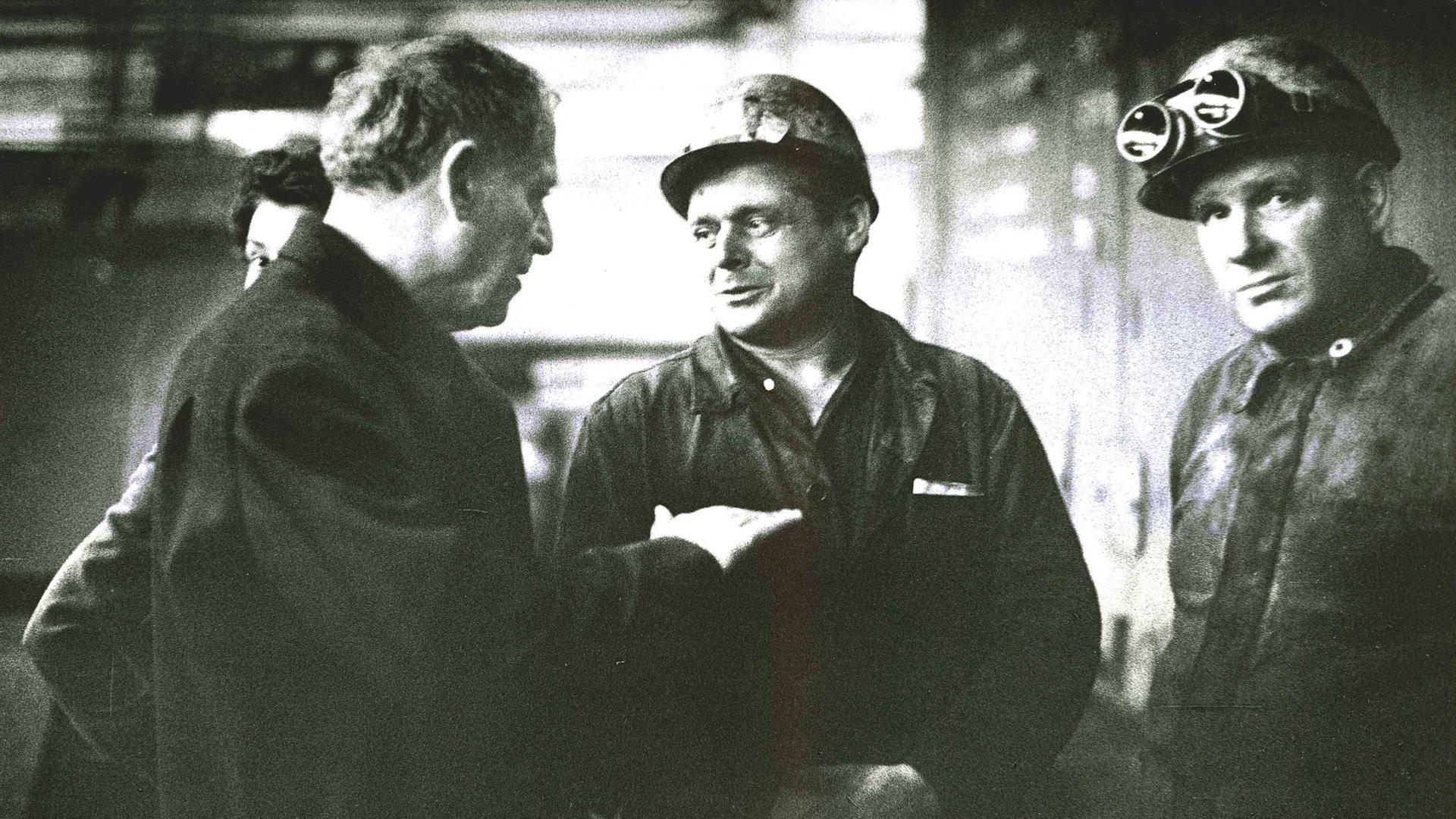 Paul Dessau in langem Mantel spricht mit zwei Grubenarbeitern in Arbeitskluft mit Helm und Schutzbrille.