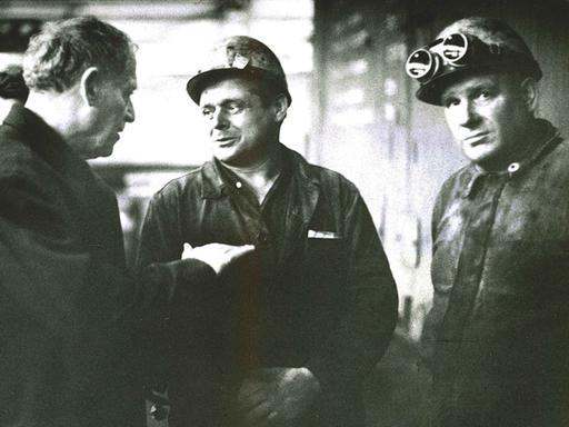 Paul Dessau in langem Mantel spricht mit zwei Grubenarbeitern in Arbeitskluft mit Helm und Schutzbrille.