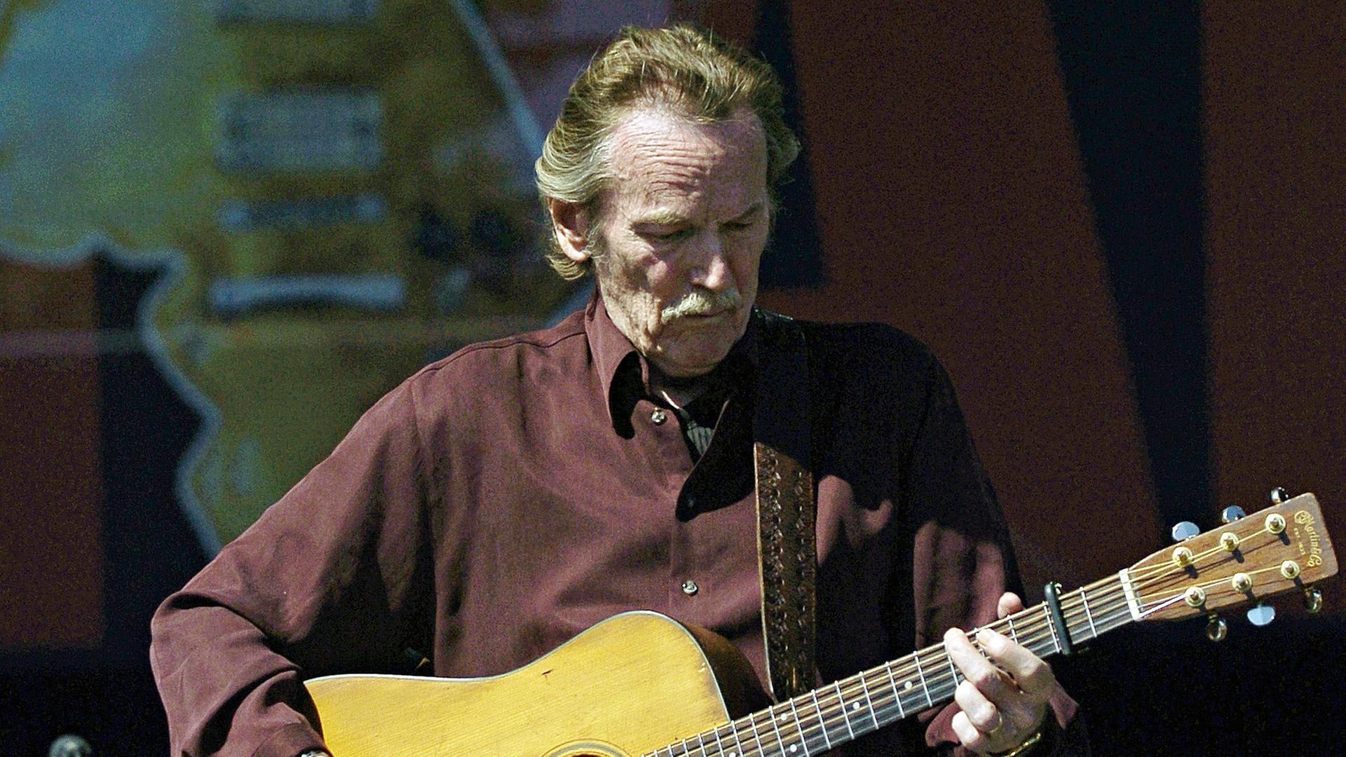 Lightfoot im dunkelroten Hemd und mit Schnurrbart spielt auf einer Bühne auf einer akustischen Gitarre.