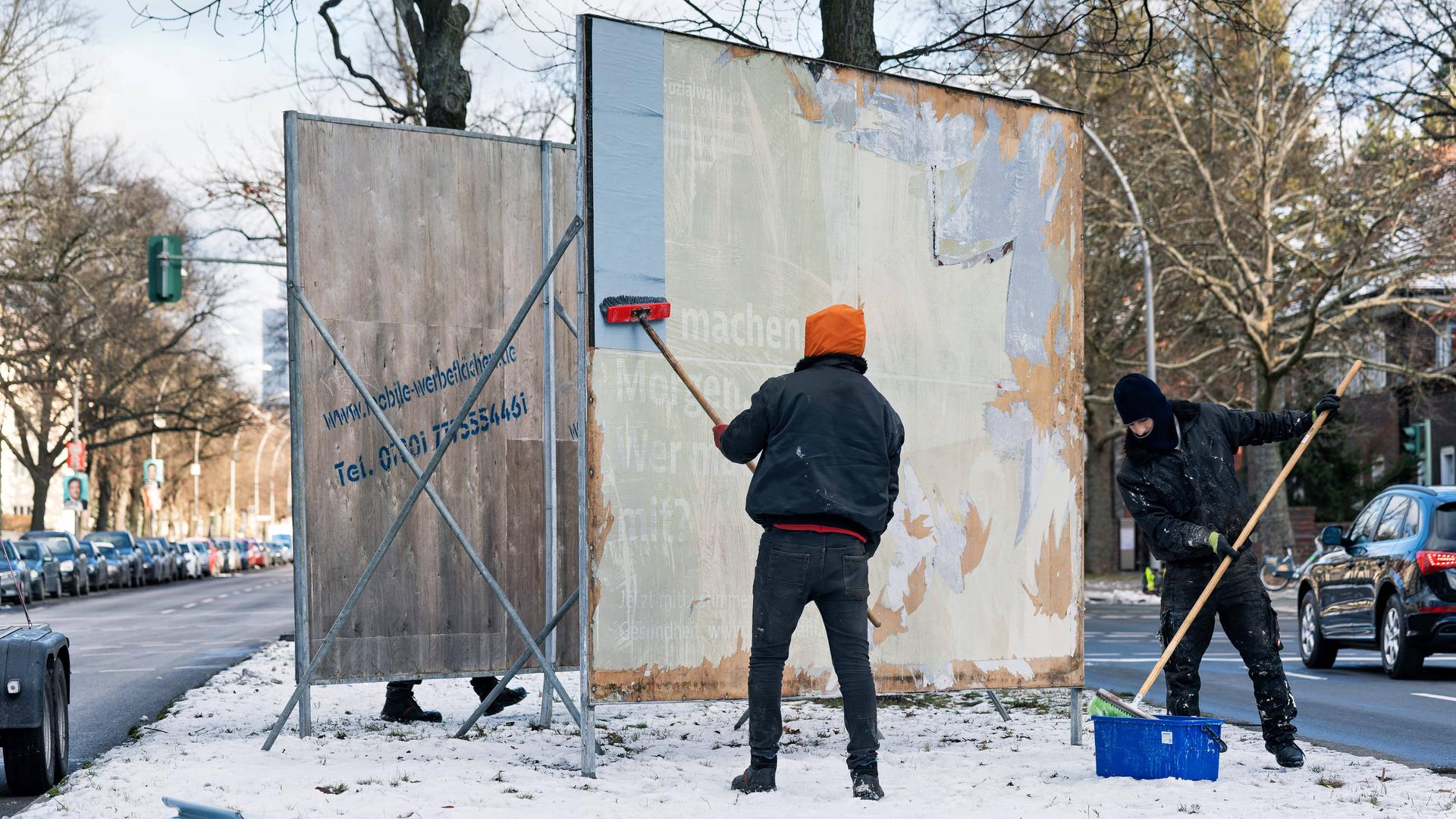 Zwei Männer mit bereiten eine leere Plakatwand auf einem verschneiten Straßenmittelstreifen vor für ein Wahlkampf-Plakat.