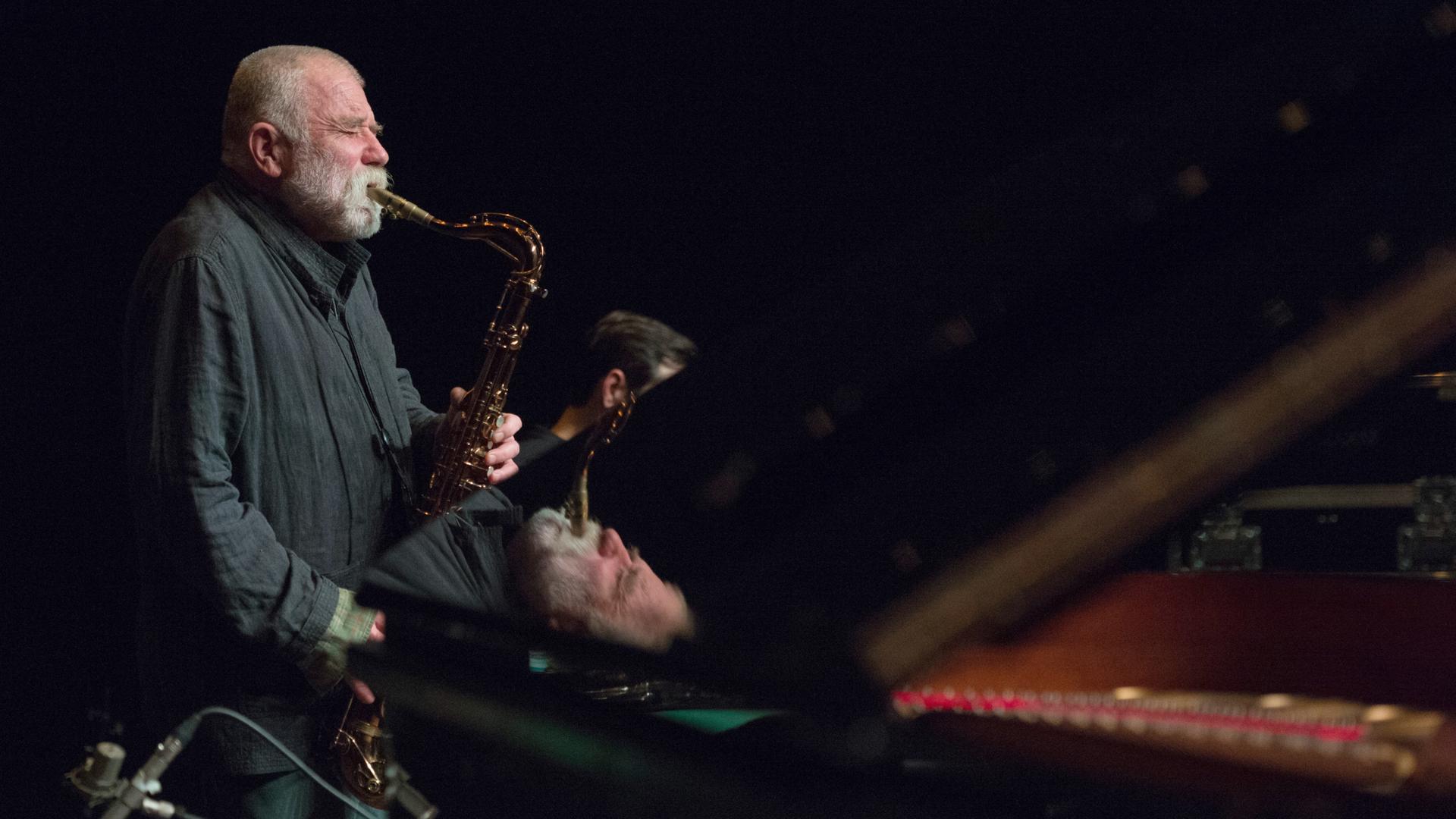 Der deutsche Jazzmusiker Peter Brötzmann auf einer dunklen Bühne und spielt Saxophon. Teile von ihm spiegeln sich in dem Flügel, der vor ihm auf der Bühne steht.