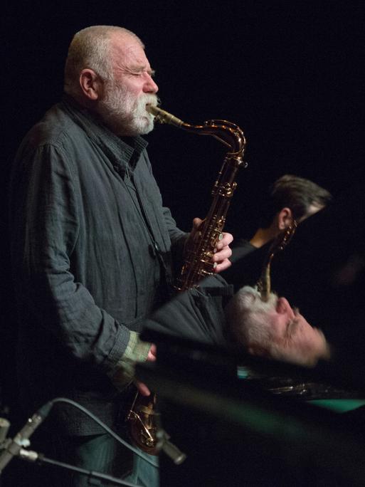 Der deutsche Jazzmusiker Peter Brötzmann auf einer dunklen Bühne und spielt Saxophon. Teile von ihm spiegeln sich in dem Flügel, der vor ihm auf der Bühne steht.