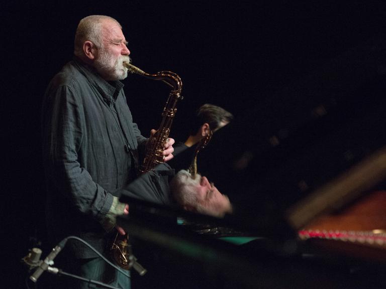 Der deutsche Jazzmusiker Peter Brötzmann auf einer dunklen Bühne und spielt Saxophon. Teile von ihm spiegeln sich in dem Flügel, der vor ihm auf der Bühne steht.