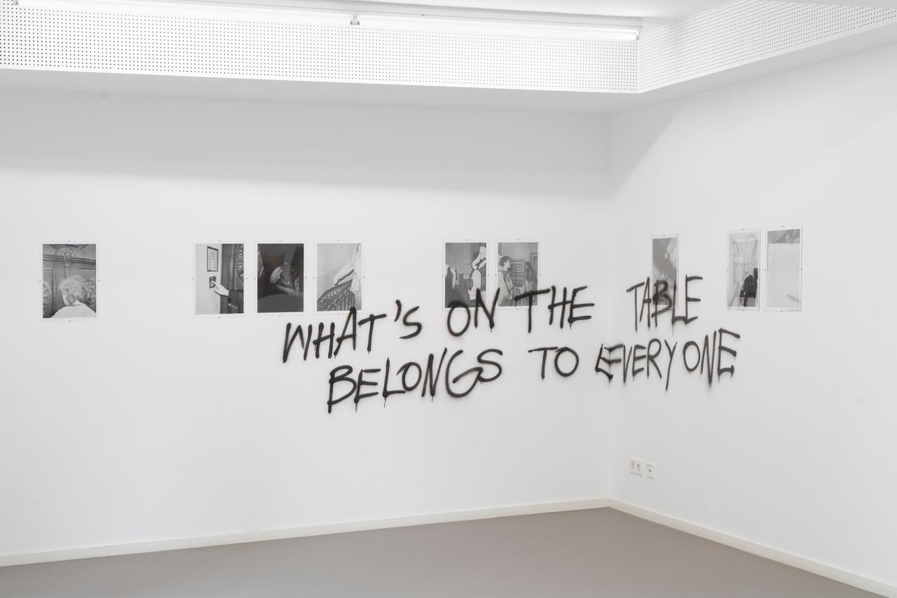 Eine Wand mit Bildern in einer Ausstellung, an der Wand schwarz getüncht der Satz "What's on the table belongs to everyone"