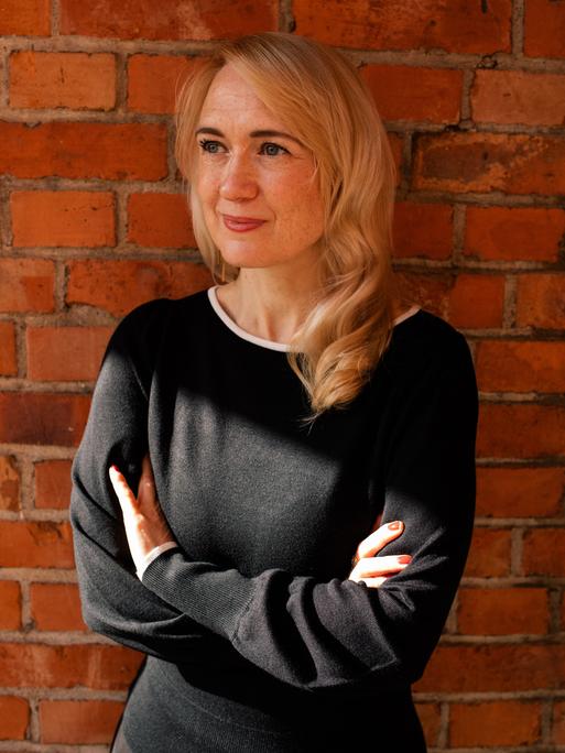 Schriftstellerin Anousch Mueller steht lächelnd vor einer Backsteinmauer. Schriftstellerin Anousch Mueller steht lächelnd vor einer Backsteinmauer.