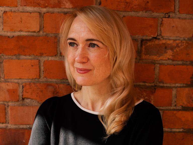 Schriftstellerin Anousch Mueller steht lächelnd vor einer Backsteinmauer. Schriftstellerin Anousch Mueller steht lächelnd vor einer Backsteinmauer.