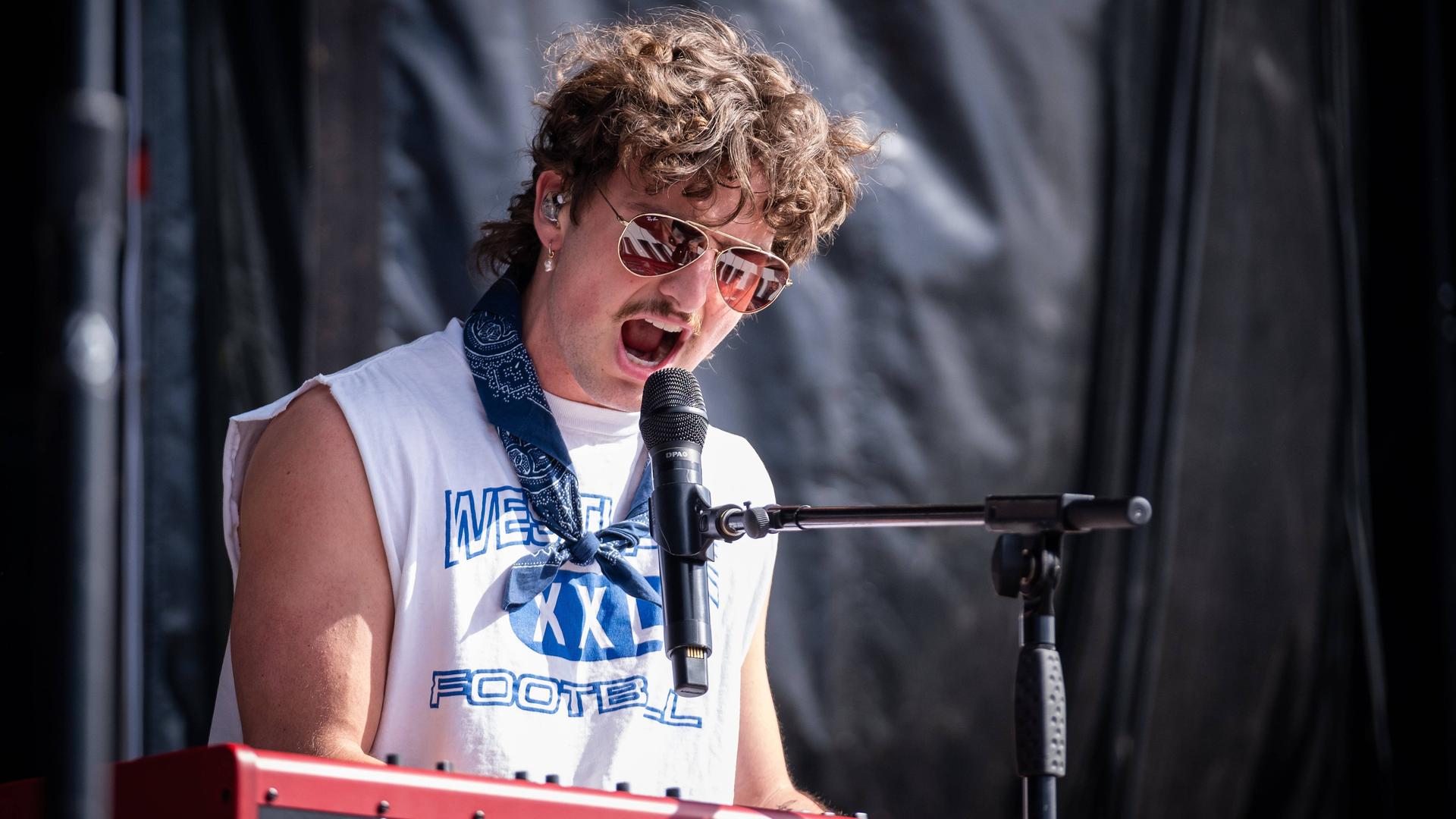 Der US-amerikanische Singer-Songwriter Benson Boone steht auf der Bühne an einem Keyboard und singt. 