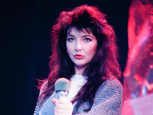 Eine Frau mit dunklen, langen Haaren hält ein Mikrofon. Durch die Beleuchtung hintrelässt ihr Haar an den Konturen einen rötlichen Eindruck.