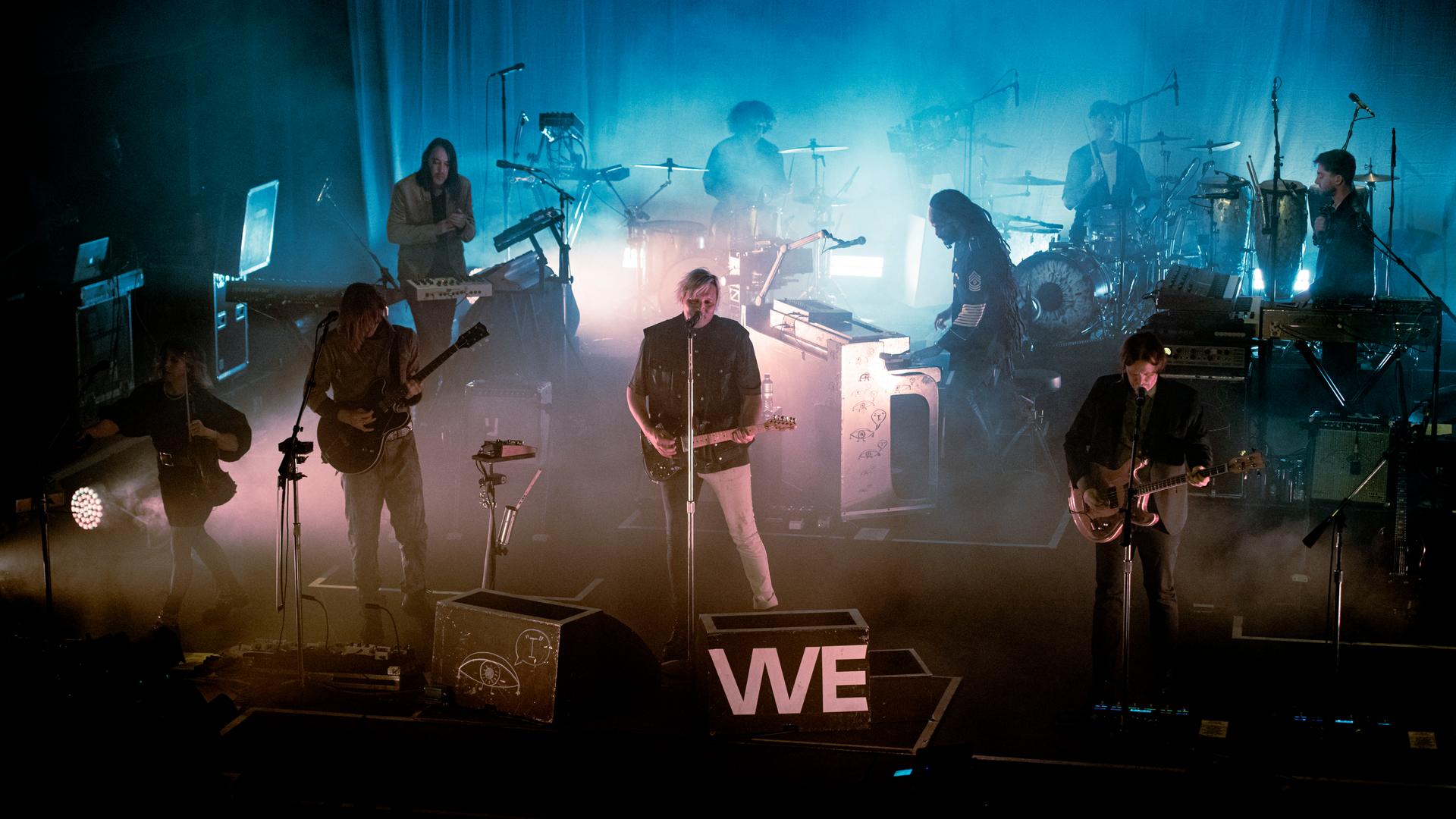 Die kanadische Indie-Band Arcade Fire bei einem Konzert in London im April 2022. Die Bühne ist dunkel, einige Lichter scheinen durch den Nebel. Am Anfang der Bühne steht in großen Buchstaben das Wort "We". Das Bild ist leicht von oben aufgenommen. 