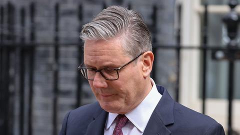 Keir Starmer steht im Anzug mit gesenktem Blick vor einem Metallzaun