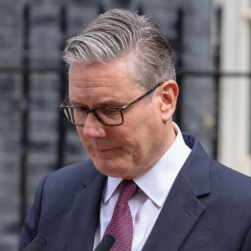 Keir Starmer steht im Anzug mit gesenktem Blick vor einem Metallzaun
