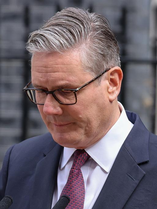 Keir Starmer steht im Anzug mit gesenktem Blick vor einem Metallzaun